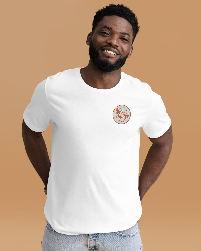 Camiseta_Accesorio_Tienda En esta imagen se muestra un hombre con una camiseta blanca que tiene el logo de la marca en la parte derecha. La imagen tiene un fondo anaranjado