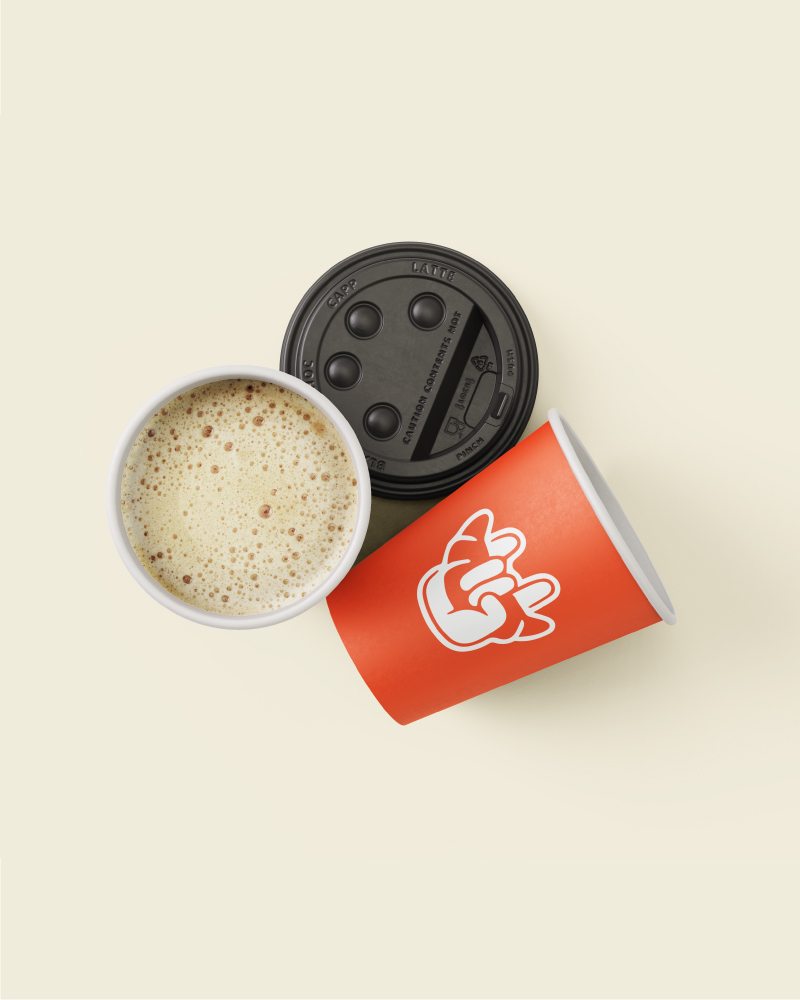 Indicación_bebidas_Apartado_Tienda Esta imagen muestra un vaso de café de color naranja con el logo de la marca y un fondo de color beis